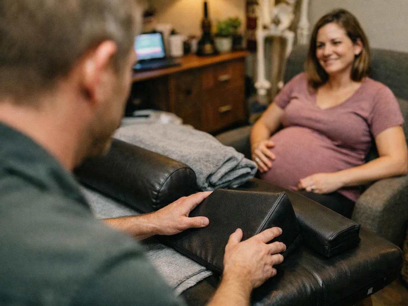 A man adjusts a black pregnancy support wedge as a smiling pregnant woman relaxes in a living room