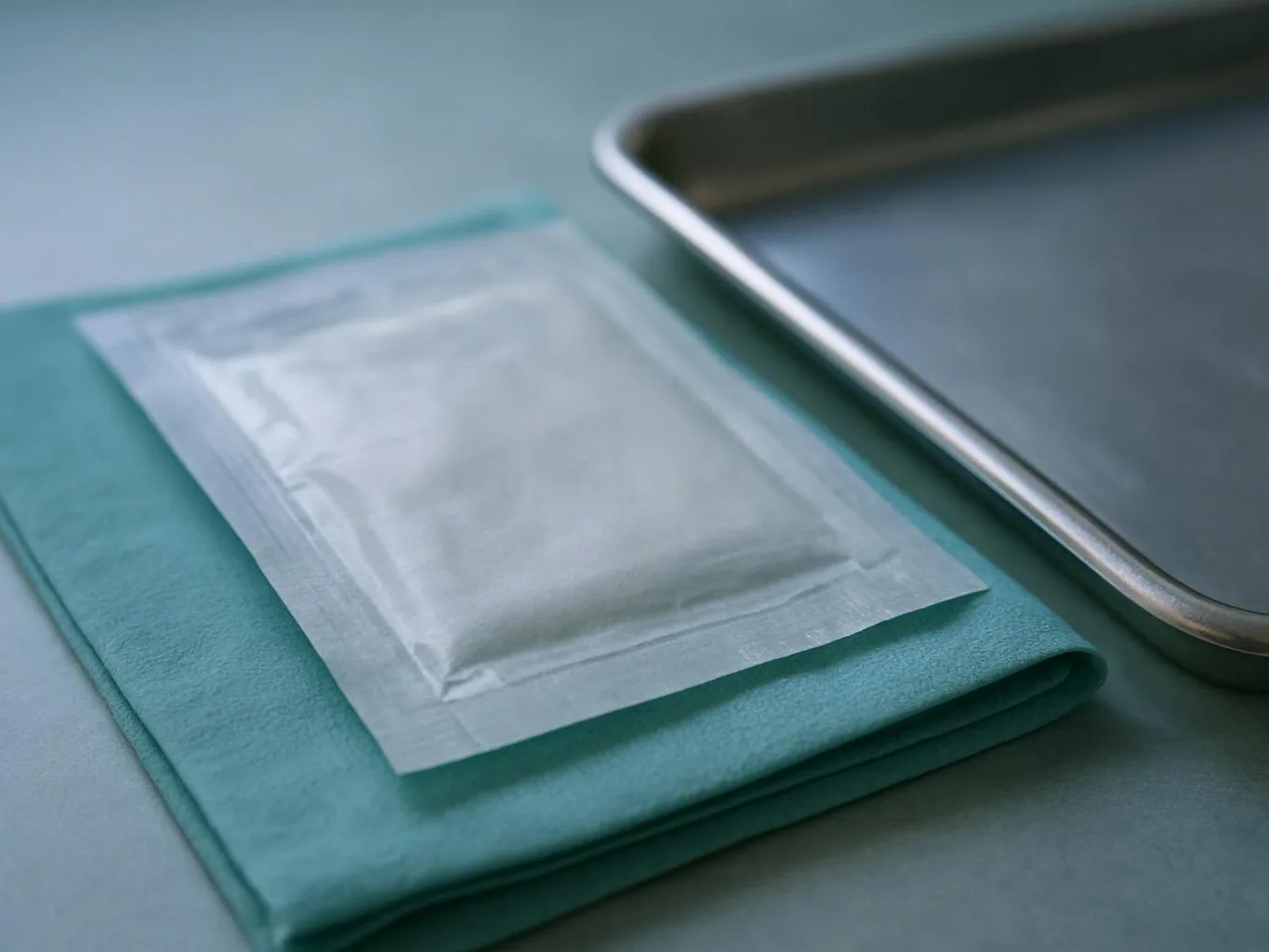 A sealed medical packet rests on a folded teal cloth beside a metal tray